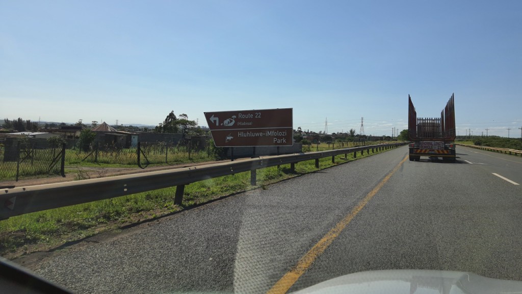 Blick aus dem Auto auf die Schnellstraße mit Schild zum Hluhluwe-iMfolozi Park (© Cornelia Hebrank, 2024)